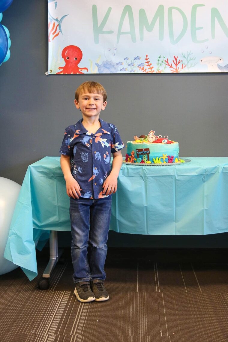 A boy smiling standing next to his birthday cake. 