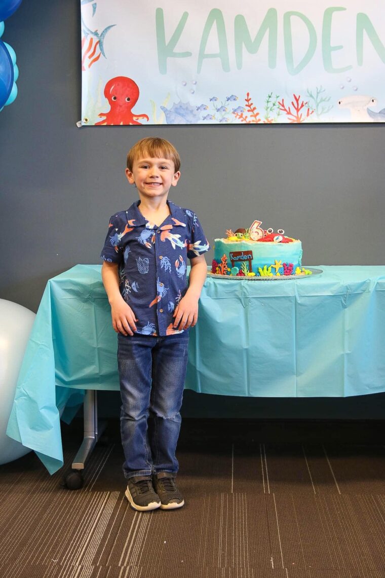 A boy smiling standing next to his birthday cake. 